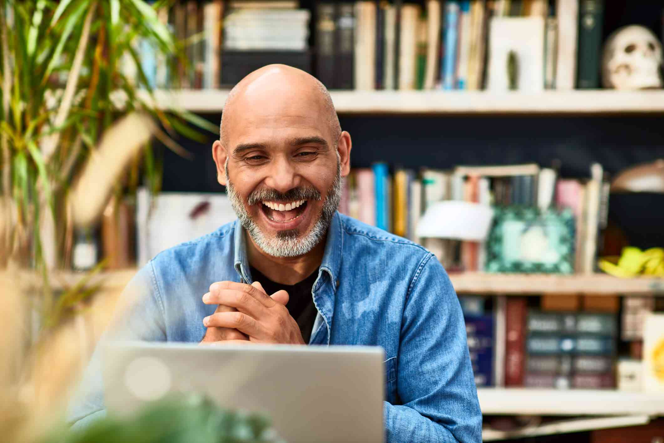 A man wearing a denim button-down shirt smiles widely and grasps his hands together as he attends an online therapy session from his home office. 